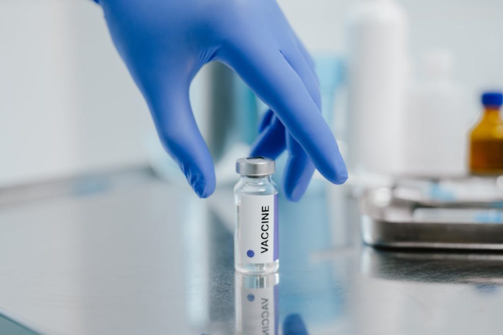 Close-up of a vaccine vial on a metal surface with a gloved hand in a laboratory setting.